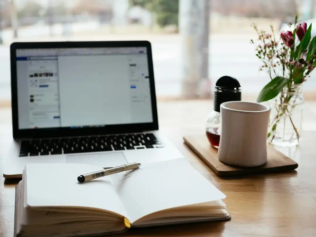 An open notebook, laptop, and cup beside a sunlit window